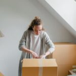 Attic Home - Slim female in cardigan unpacking cardboard box put on bed close to suitcase while standing near window in attic style room in daylight