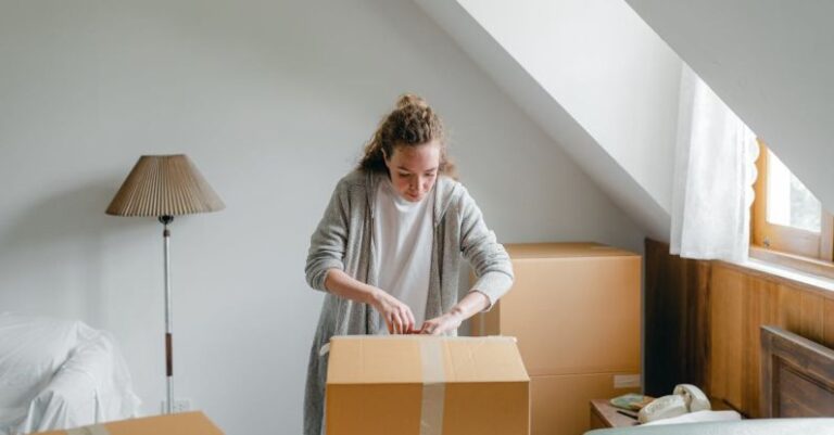 Attic Home - Slim female in cardigan unpacking cardboard box put on bed close to suitcase while standing near window in attic style room in daylight