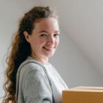 Attic Home - Side view of cheerful female in cardigan with wavy hair holding big cardboard box and looking at camera in attic style living room