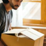 Attic Home - Side view of pensive ethnic male in casual wear and smart watch looking away while taking cardboard box from bed and standing near window after moving into new house