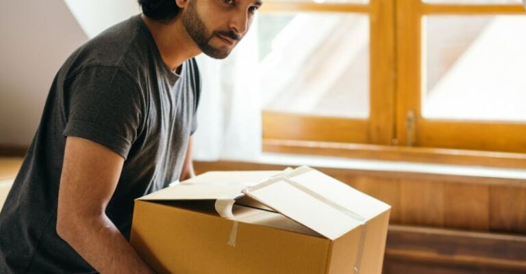 Attic Home - Side view of pensive ethnic male in casual wear and smart watch looking away while taking cardboard box from bed and standing near window after moving into new house