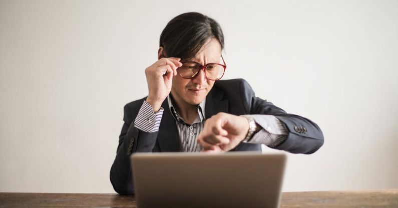 Meet Deadlines - Young frowning man in suit and glasses looking at wristwatch while waiting for appointment sitting at desk with laptop