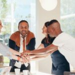 Structural Integrity - Content multiethnic colleagues joining hands above table with laptops in modern office on sunny day